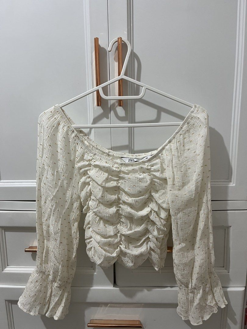 Zara White Ruched Square Neck Long Sleeve Top (Category: Blouses) located in Pili. Submitted by jhamimin22 (Listing ID 1462149507, Image 1). Description: - White Zara top - Ruched front detail - Elegant square neckline - Long sleeves with ruffled cuffs - Subtle gold dot pattern throughout - Convenient side zipper closure - Versatile for dressing up or down.
