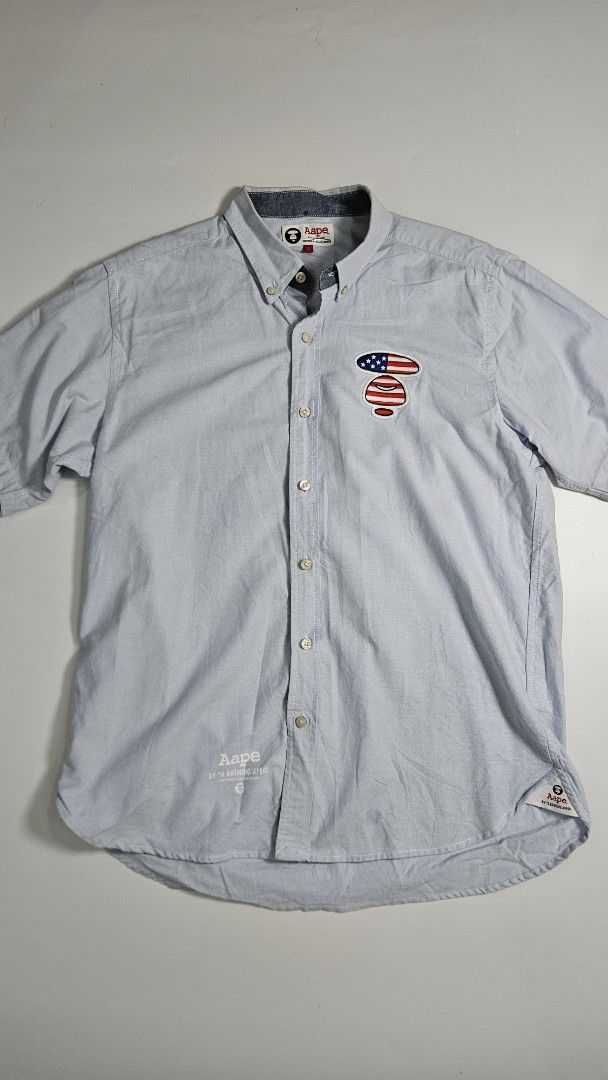 Aape by A Bathing Ape Blue Button Down Shirt (Category: Tshirts & Polo Shirts ) located in Alfonso. Submitted by jbcamet (Listing ID 1462415519, Image 1). Description: - Brand: Aape by A Bathing Ape - Color: Light Blue - Material: 100% Cotton - Design: Button-down shirt - Features: Embroidered American flag patch on chest, Aape logo print on hem - Collar: Classic button-down - Cuffs: Button cuffs.