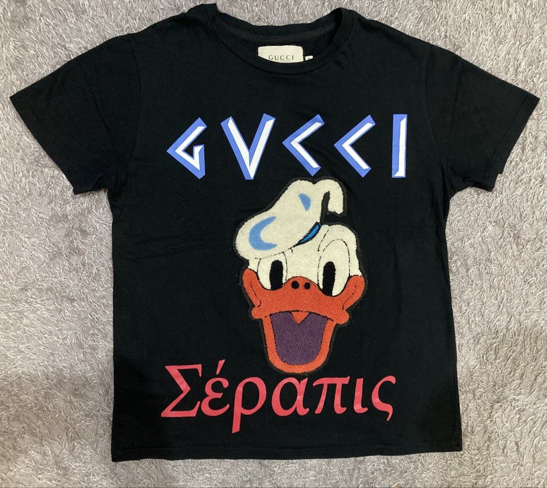 Gucci tshirt (Category: Luxury Apparel) located in General Santos City (Dadiangas). Submitted by amorhesthrift (Listing ID 1462381347, Image 1). Description: Like new condition Size-Medium on tag Dimes- W19.5 L24 This black short-sleeve T-shirt features a playful parody design combining a stylized Gucci-style logo, a fuzzy Donald Duck appliqué, and the Greek word for Serapis at the bottom.