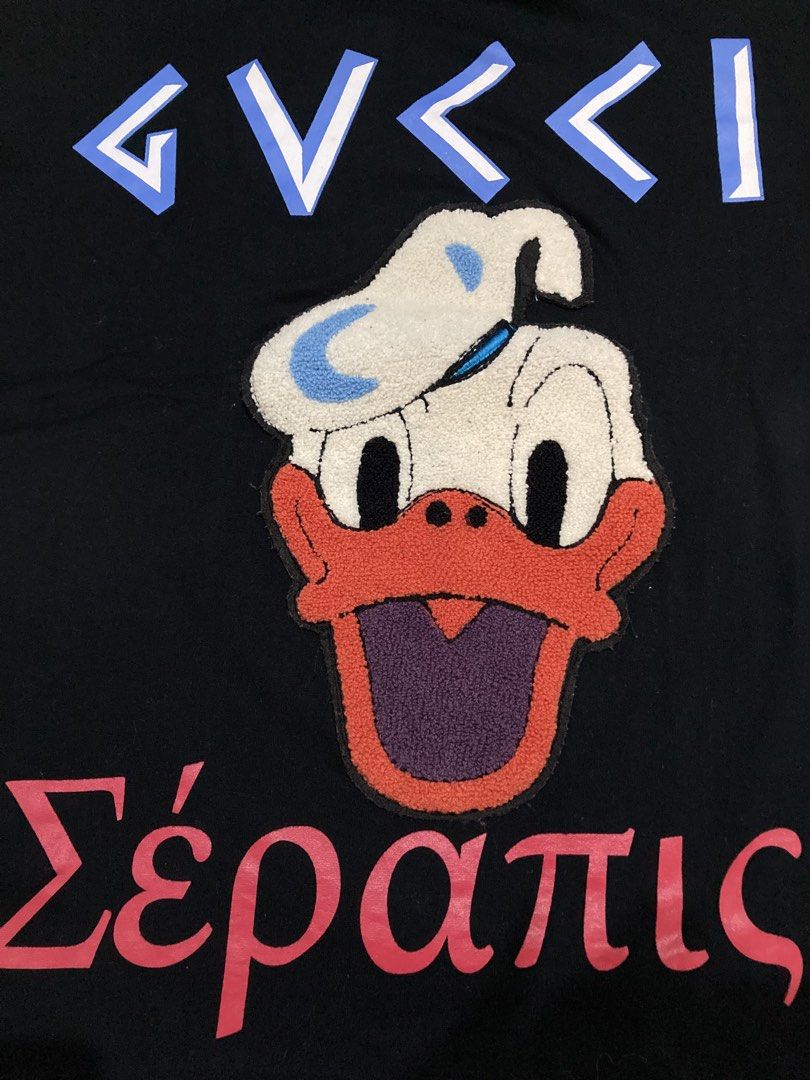 Gucci tshirt (Category: Luxury Apparel) located in General Santos City (Dadiangas). Submitted by amorhesthrift (Listing ID 1462381347, Image 2). Description: Like new condition Size-Medium on tag Dimes- W19.5 L24 This black short-sleeve T-shirt features a playful parody design combining a stylized Gucci-style logo, a fuzzy Donald Duck appliqué, and the Greek word for Serapis at the bottom.