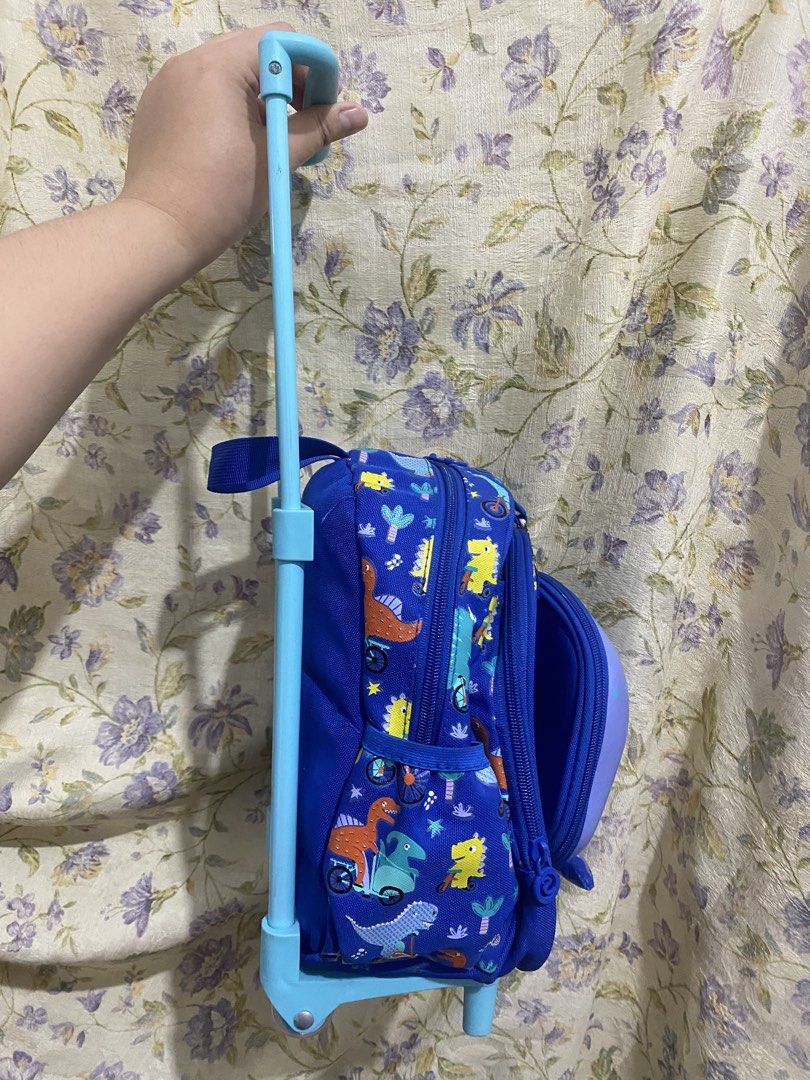 Kids Blue Dinosaur Rolling Backpack with Wheels (Category: Backpacks) located in Binan City. Submitted by prelovedlagunaph (Listing ID 1462620455, Image 3). Description: - Adorable blue rolling backpack for kids. - Features a fun all-over print of dinosaurs and vehicles. - Cute purple whale face design on the front. - Includes a plush toy hanging from the whale face. - Extendable handle for easy pulling and maneuverability. - Ideal for school, da