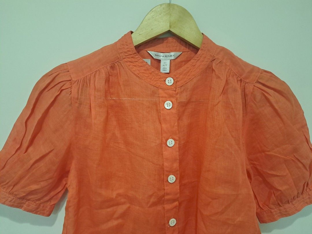 Banana Republic linen fabric (Category: Blouses) located in Manila. Submitted by joanbalde39643 (Listing ID 1462798309, Image 2). Description: This is a women's Banana Republic short-sleeve button-up blouse crafted from 100% ramie or linen. It features a vibrant coral orange tone, a classic band/mandarin collar, white contrast buttons down the front, and feminine puff sleeves..