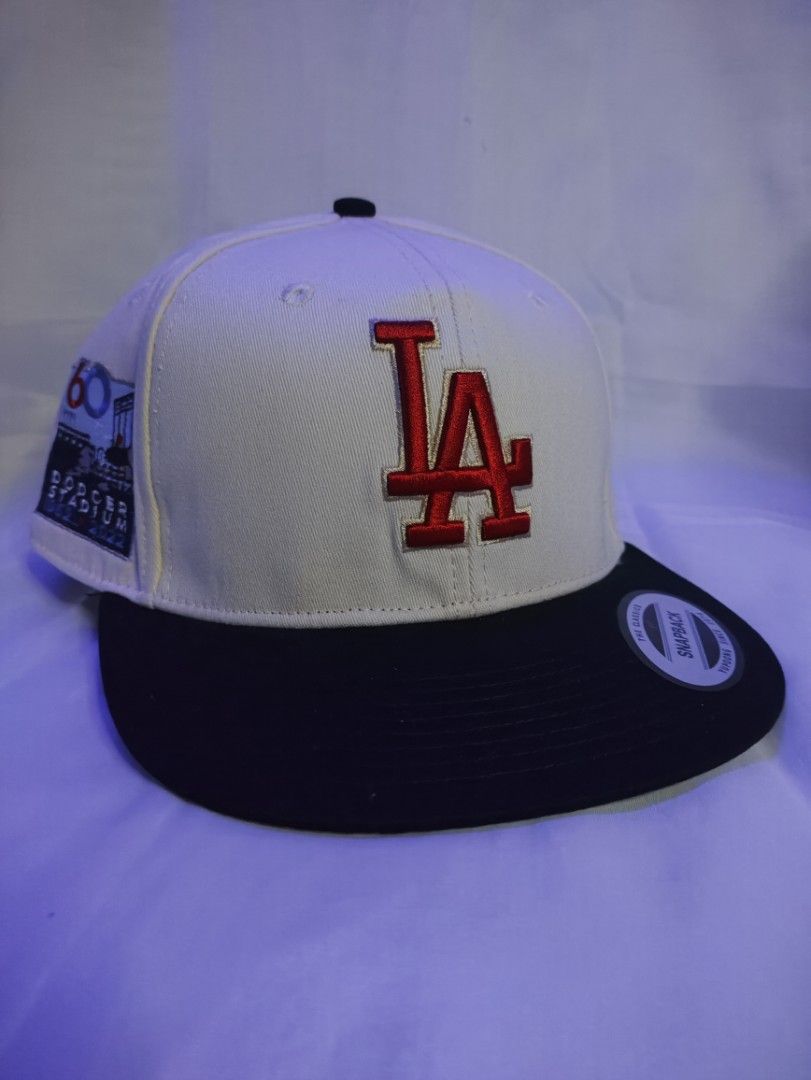 LA Dodgers Snapback Cap - Dodger Stadium 60th Anniversary (Category: Caps & Hats) located in Manila. Submitted by vestige_collection (Listing ID 1462863979, Image 1). Description: - LA Dodgers snapback hat - Cream colored crown with red 'LA' logo - Black brim - Special patch: Dodger Stadium 60th Anniversary (1962-2022) - MLB logo on the back - Adjustable snapback closure - Size: 7 1/8.