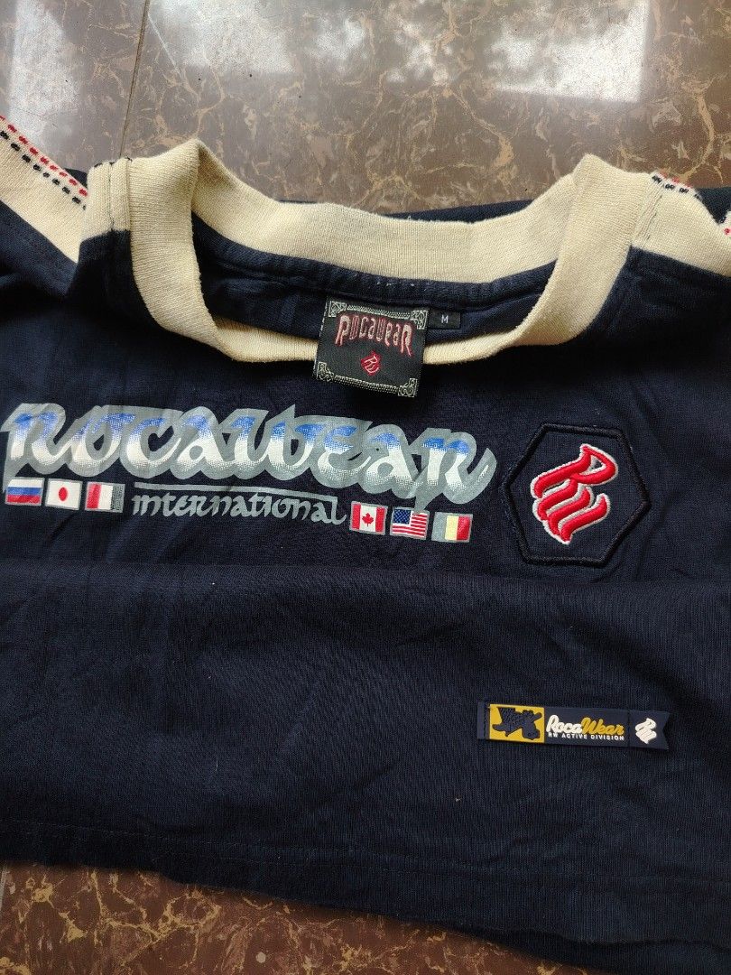 Rocawear International T-Shirt (Category: Tshirts & Polo Shirts ) located in Alegria. Submitted by goofy_ripper (Listing ID 1462760592, Image 2). Description: - Navy blue Rocawear t-shirt - Features 'Rocawear International' graphic with flags - Rocawear logo on the sleeve - Made of 100% cotton - Size: Medium (M) - Ideal for casual wear.