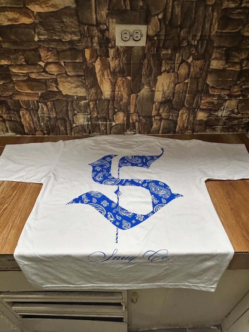 SNUG CO. White T-Shirt (Category: Tshirts & Polo Shirts ) located in Cavite. Submitted by nunoy_08 (Listing ID 1462932626, Image 2). Description: - Brand: SNUG CO. - Color: White - Design: Bold blue paisley graphic on front and back - Front Detail: 'SNUG CO.' in large letters - Back Detail: Large 'S' with 'Snug Co.' below - Material: Good quality cotton - Occasion: Great for casual wear.