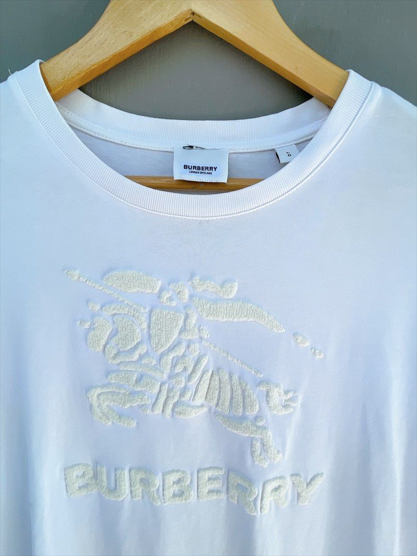 Burberry White Embossed Logo T-Shirt (Category: Tshirts & Polo Shirts ) located in San Nicolas. Submitted by .luxfinds_0222 (Listing ID 1463319369, Image 2). Description: Size: L-XL Width :23 Length :28 CR:9.5 Issue minimal stain/repair pinhole - Burberry T-shirt - Size L (based on tag) - Features embossed Horseferry print and logo - Short sleeves, crew neck - Small repair pinhole on front (refer to last photo).