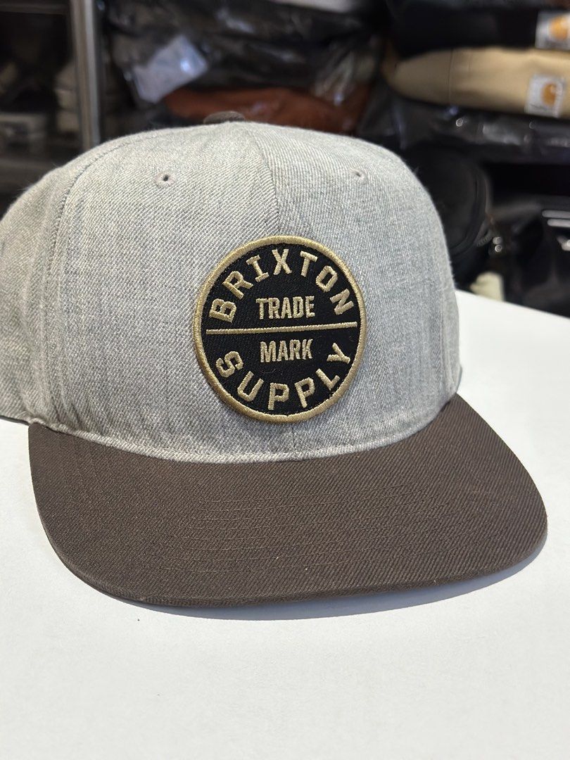 Brixton Trade Mark Supply Snapback Hat (Category: Caps & Hats) located in Daet. Submitted by thrifthings2018 (Listing ID 1463425275, Image 2). Description: - Brand: Brixton - Style: Snapback hat - Colors: Grey and brown - Features: 'Brixton Trade Mark Supply' logo patch on front - Fit: Adjustable strap at the back - Design: Classic flat brim - Ideal for: Everyday wear.