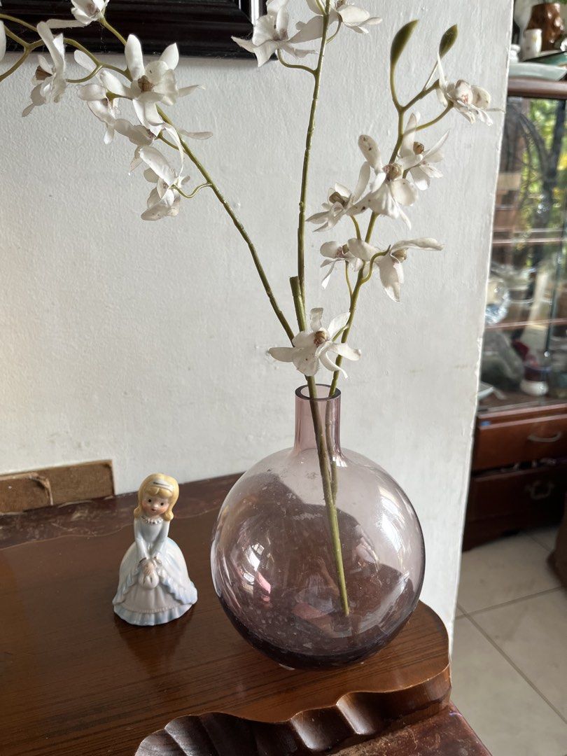Ikea hand blown Glass Vase (Category: Vases & Decorative Bowls) located in Taguig. Submitted by minmin_parker (Listing ID 1463430507, Image 2). Description: - Elegant 8-inch tall round glass vase with a subtle purple hue. - Comes with realistic artificial white orchid stems. - Includes a charming small porcelain doll figurine. - Ideal for enhancing shelf or table decor. - Makes a thoughtful gift for any occasion..