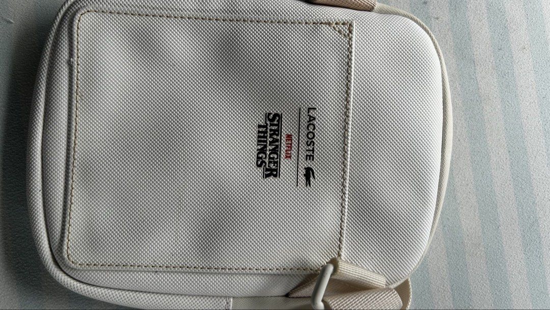 Lacoste x Stranger Things Crossbody Bag White (Category: Cross-body Bags) located in Bulacan. Submitted by peachyaguilar (Listing ID 1463381168, Image 2). Description: - White Lacoste crossbody bag - Features a Stranger Things graphic on the front pocket - Zipper closure - Adjustable strap - Ideal for everyday use or travel - Small and compact design.