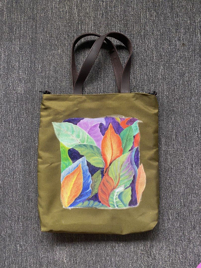Olive Green Tote Bag with Hand-Painted Design (Category: Tote Bags) located in Makati City. Submitted by clutterfree.life (Listing ID 1463389124, Image 1). Description: - Stylish olive green tote bag - Vibrant, colorful leaf illustration on the front - Durable brown leather handles - Secure zippered main compartment - Convenient exterior pockets on the back - Black nylon fabric interior lining - Ideal for everyday use or as a fashion statement On