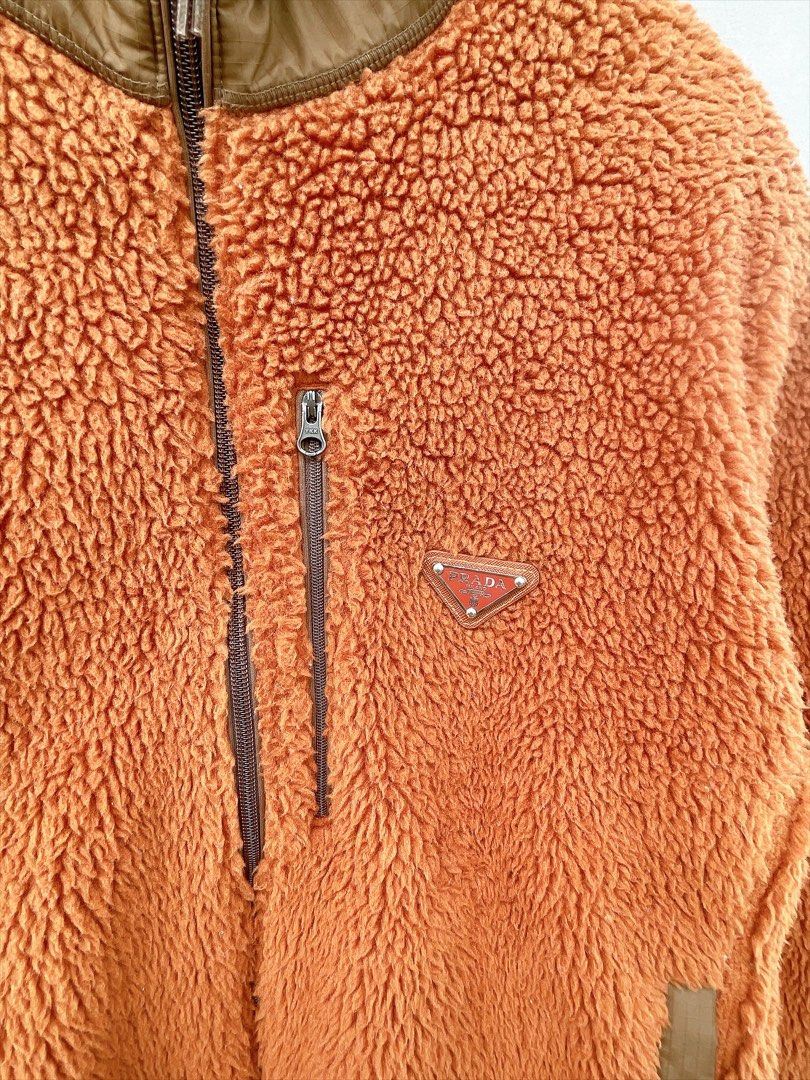 Prada Orange Fleece Zip-Up Jacket (Category: Luxury Apparel) located in San Nicolas. Submitted by .luxfinds_0222 (Listing ID 1463333319, Image 2). Description: Size: L-XL Width :24 Length :28 CR:10/10 Issue :NONE - Cozy brown fleece jacket - Brown trim details - Zip-up front closure - Chest pocket with authentic Prada logo - Two convenient side pockets - Durable YKK zippers - Ideal for warmth and layering during cooler seasons.