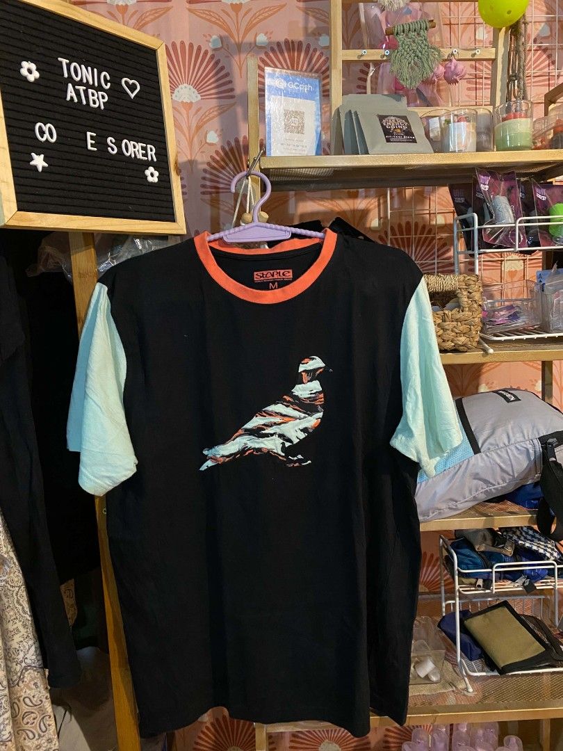 Staple "Pigeon " tee (Category: Tshirts & Polo Shirts ) located in Aroroy. Submitted by dr0p-a-dr1p (Listing ID 1463431230, Image 1). Description: - Brand: Staple - Color: Black with light blue sleeves and orange collar - Graphic: Unique abstract pigeon design in grey, black, and orange - Material: 100% Cotton - Size: Medium (M) - Style: Casual wear.