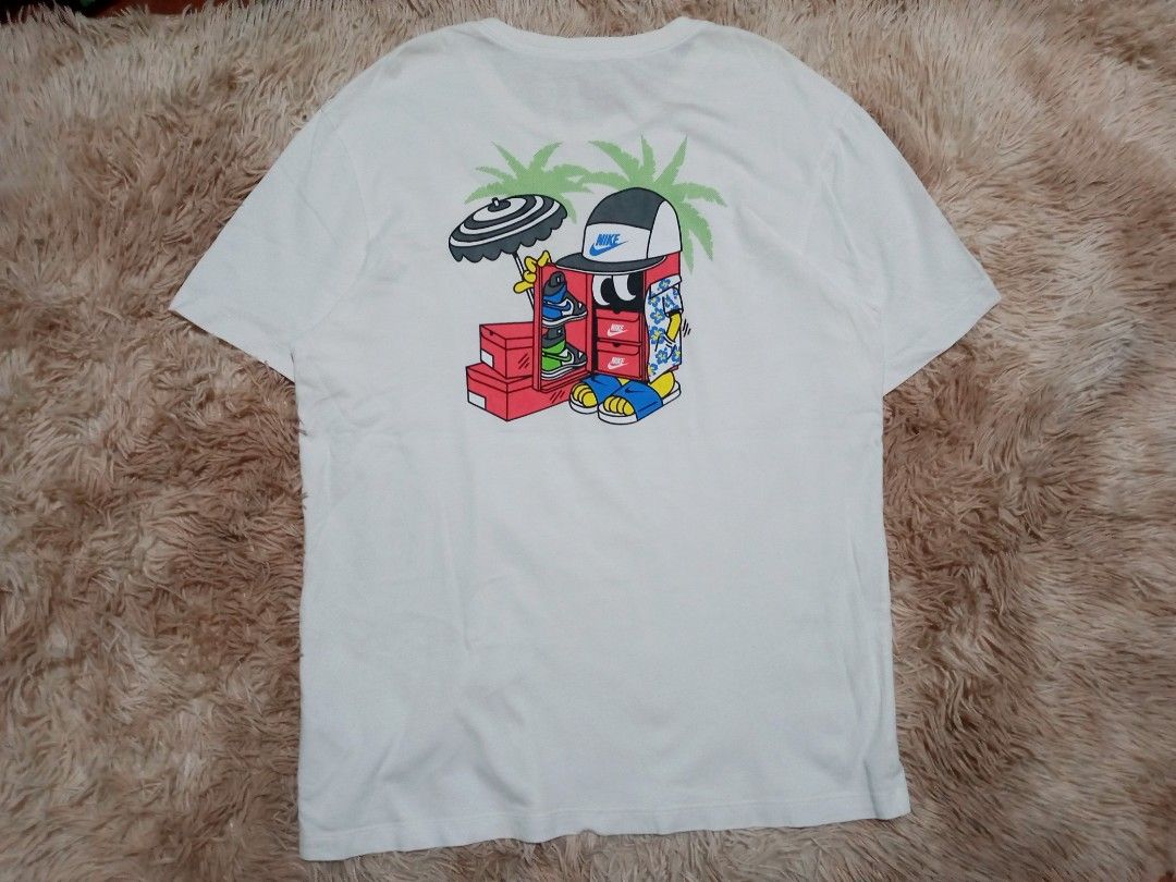 Nike Tee T-Shirt White Graphic Print (Category: Activewear) located in Boston. Submitted by johnpaulvi30029 (Listing ID 1463681769, Image 1). Description: - White Nike T-shirt - Features a fun graphic on the back with a cartoon character, sneakers, and palm trees - Small graphic on the front chest area - Nike Tee label visible on the collar - Ideal for casual wear.