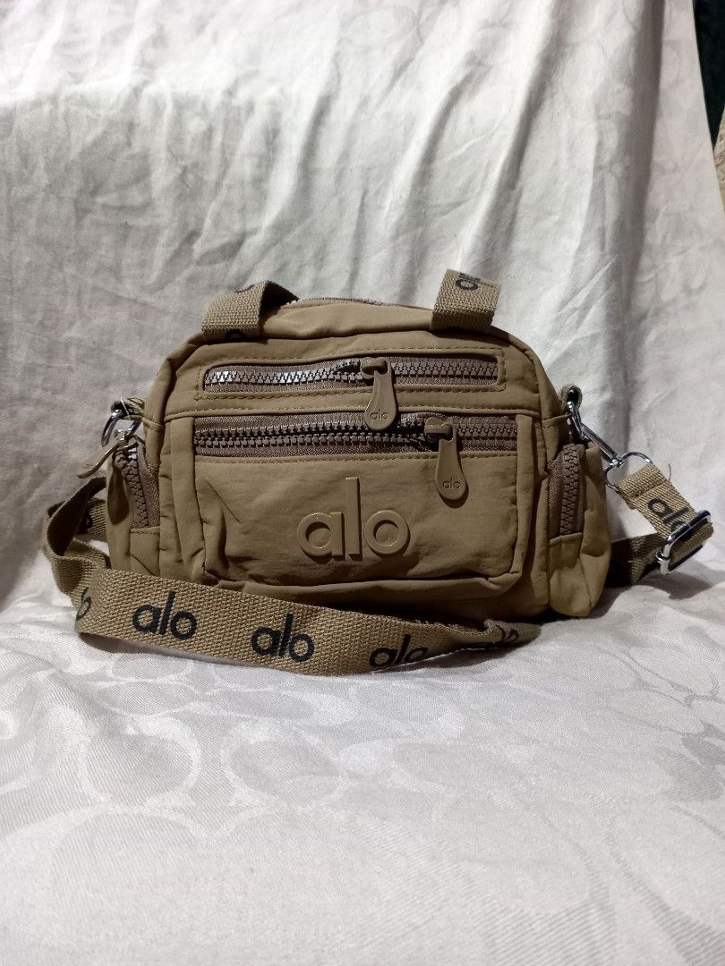 Alo Yoga crossbody bag (Category: Cross-body Bags) located in Aguinaldo. Submitted by ukayandthrift (Listing ID 1463768507, Image 1). Description: - Tan Alo Yoga crossbody bag - Multiple zippered compartments for secure storage - Adjustable and detachable strap with repeating Alo logo - Versatile for everyday use or travel.