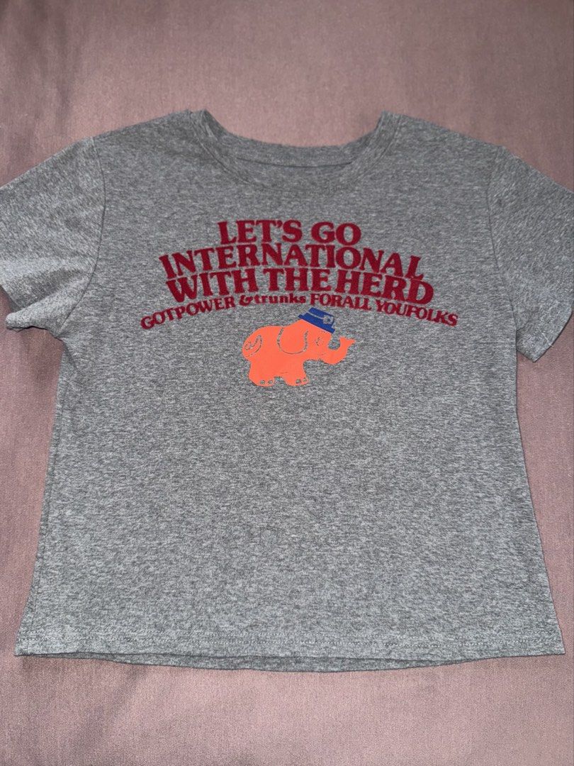 Baby Tee Grey with Embossed Elephant Design (Category: Shirts) located in Binan City. Submitted by andis_collection (Listing ID 1463999397, Image 1). Description: - Grey t-shirt with a unique graphic design. - Front text: 'LET'S GO INTERNATIONAL WITH THE HERD' and an orange elephant graphic. - Back text: 'ELEPHANTS WILL TRAVEL' and 'GENTLE FEELINGS & COMPASSION'. - Features embossed details for a premium touch. - Ideal for everyday casual wear. -