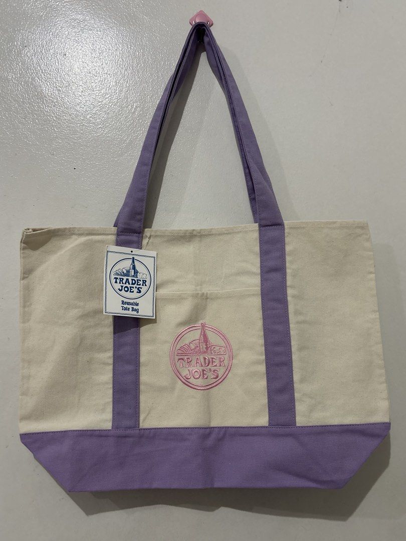 Trader Joe's Reusable Tote Bag (Large) (Category: Tote Bags) located in Baler. Submitted by HelpDeclutterMyCloset (Listing ID 1464071027, Image 1). Description: - Authentic Trader Joe's reusable tote bag - Stylish cream color with vibrant purple accents and handles - Features the iconic Trader Joe's logo embroidered on the front - Brand new with original tags attached - Ideal for grocery shopping or everyday errands.