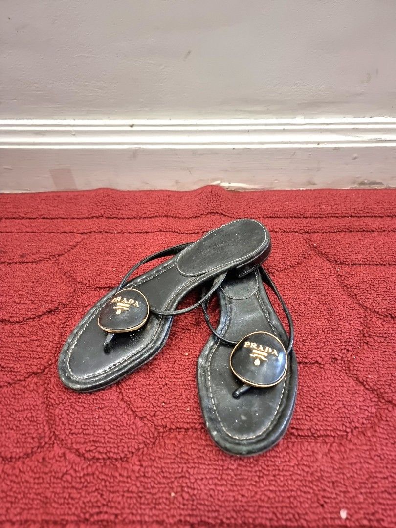 Prada Black Leather Thong Sandals with Gold Logo (Category: Flats & Sandals) located in Manila. Submitted by pickmemanila (Listing ID 1464147985, Image 1). Description: - Authentic Prada black leather thong sandals - Features iconic gold Prada logo on front strap - Comfortable flat sole - Versatile for casual or dressing up outfits - Shows some signs of wear on soles and straps, but plenty of life left - Classic designer piece.