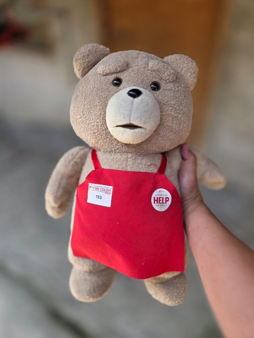 Ted Plush Toy with Red Apron and "Help" Button (Category: Plushies & Soft Toys) located in Santa Maria. Submitted by purefalcon-f7cf (Listing ID 1464362341, Image 2). Description: - Cute Ted plush toy - Original tag still attached - Wearing a red apron with 'Bay Colony Today' name tag - Features a 'How May We Help You Today?' button - Red ribbon tied in a bow on the back - Great for Ted movie fans or as a collectible.