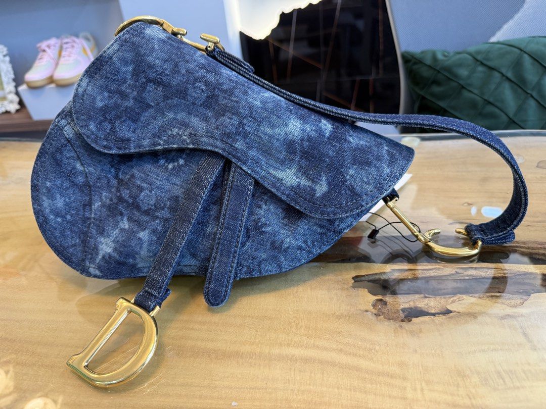 Christian Dior Denim Saddle Bag (Category: Luxury Bags & Wallets) located in Alfonso. Submitted by luxurose (Listing ID 1464758927, Image 1). Description: Christian Dior saddle bag in blue denim. Distinctive curved shape with the iconic hanging 'D' charm and 'CD' hardware on the strap attachments. Tie-dye or bleach wash pattern throughout with a circular motif on the back pocket. Short shoulder strap with contrast stitching. Clean hardware and de