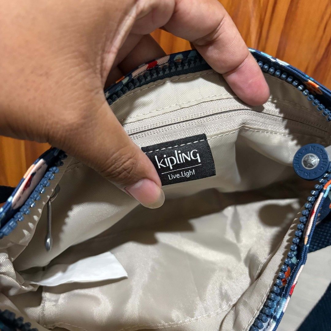 Kipling Blue Floral Crossbody Bag with Monkey Keychain (Category: Cross-body Bags) located in Carmen. Submitted by ar_thrift00 (Listing ID 1464714245, Image 3). Description: - Brand: Kipling - Style: Crossbody bag with adjustable strap and metal hardware - Color/Design: Navy blue with an all-over colorful floral pattern - Inclusion: Classic fuzzy blue monkey keychain - Interior: Beige fabric lining with a zipper pocket and official 'Live Light' c
