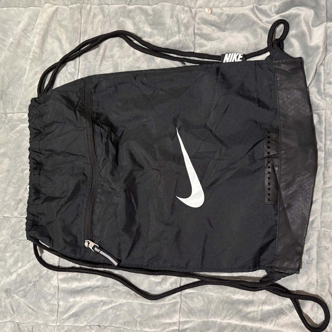 Nike Gymsack Water-Resistant Black Drawstring Backpack (Category: Backpacks) located in Carmen. Submitted by ar_thrift00 (Listing ID 1464715285, Image 3). Description: Black Nike drawstring backpack made from water-resistant fabric. Large white swoosh printed on the front. Handy front zippered pocket with a reflective pull tab. Drawstring closure with sturdy black cords. Small Nike tag attached near the top opening. Lightweight gym bag, great for