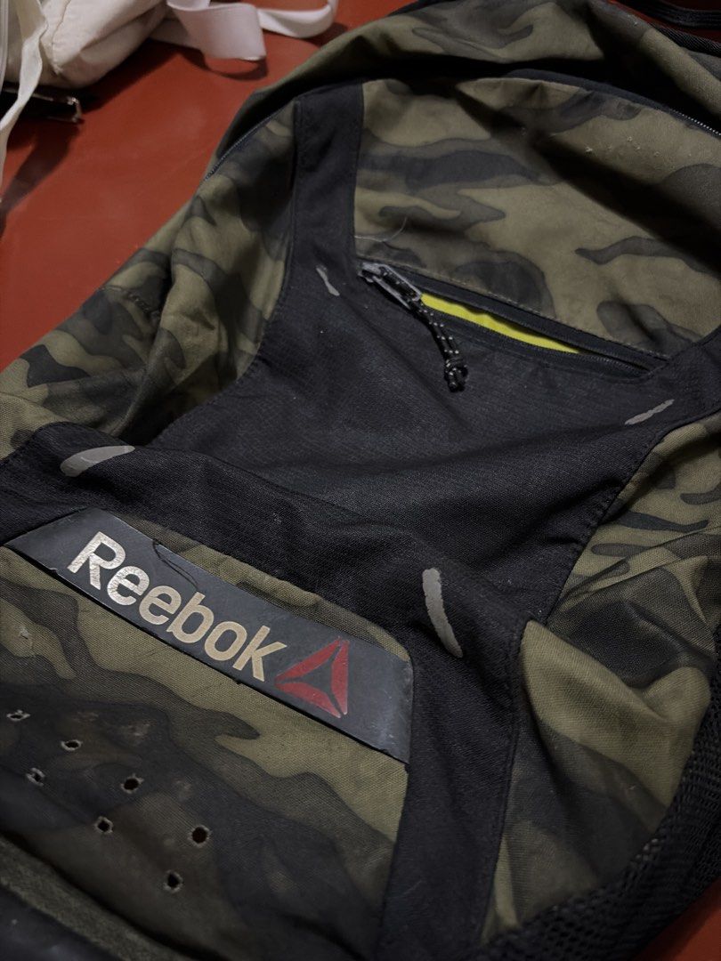 Reebok Camo Backpack (Category: Backpacks) located in Manila. Submitted by finderskeepersmnl_092026 (Listing ID 1464722206, Image 2). Description: - Authentic Reebok backpack in a stylish green camouflage print - Bold black front panel with a zippered pocket featuring a vibrant yellow interior - Signature Reebok logo patch near the bottom - Comfortable mesh shoulder straps and breathable perforation holes at the base - Spacious main compartment i