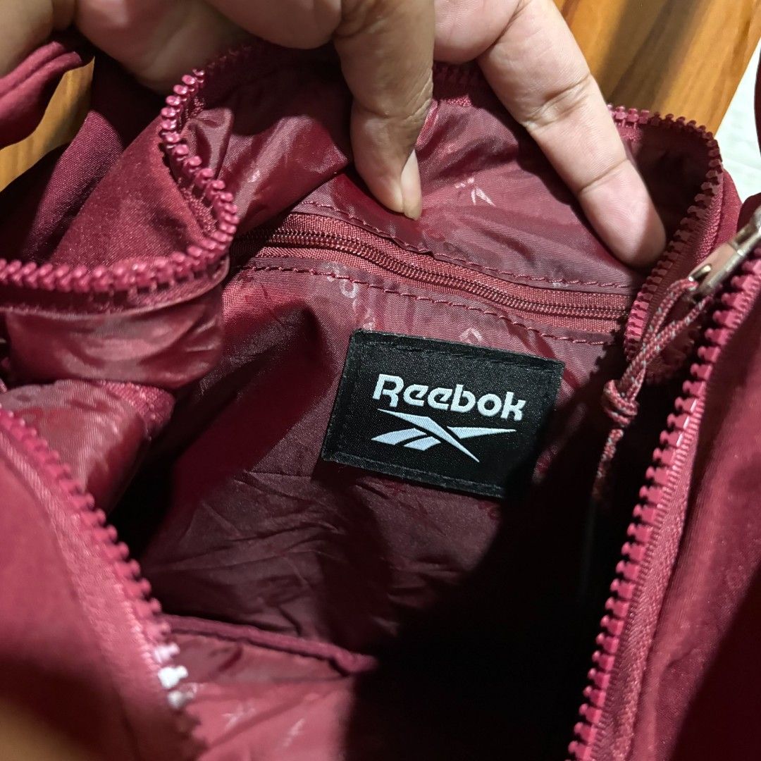 Reebok Maroon Crossbody Shoulder Bag (Category: Cross-body Bags) located in Carmen. Submitted by ar_thrift00 (Listing ID 1464711512, Image 2). Description: - Brand: Reebok - Color: Maroon with bold white front logo - Material: Lightweight nylon with deep burgundy interior lining - Strap: Adjustable shoulder strap with stylish patterned pull cord zipper detail - Storage: Main compartment plus an extra internal zipped pocket and brand tag patch - C