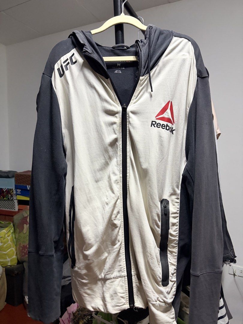 Reebok UFC Full Zip Hoodie Jacket (Category: Coats, Jackets and Outerwear) located in Manila. Submitted by finderskeepersmnl_092026 (Listing ID 1464744457, Image 1). Description: - Authentic Reebok UFC full-zip hoodie - Size: Medium (tag inside collar) - Color: White front panels with dark grey sleeves and hood - Branding: UFC logo near right shoulder and large Reebok logo on left chest - Features: Adjustable drawstring hood and secure zippered f