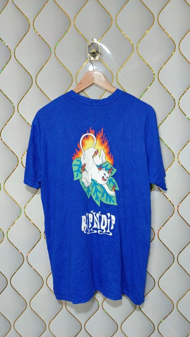 RIPNDIP Blue Graphic T-shirt (Category: Tshirts & Polo Shirts ) located in Manila. Submitted by zariyah.1986 (Listing ID 1464756143, Image 2). Description: Bright blue RIPNDIP tee with a small logo hit on the front chest. Back has a large graphic of the white cat surrounded by flames and green leaves. Standard crewneck and short sleeves. Tag says size Large, measures approximately 21 inches pit to pit by 29 inches length. Cotton fabric, soft feel