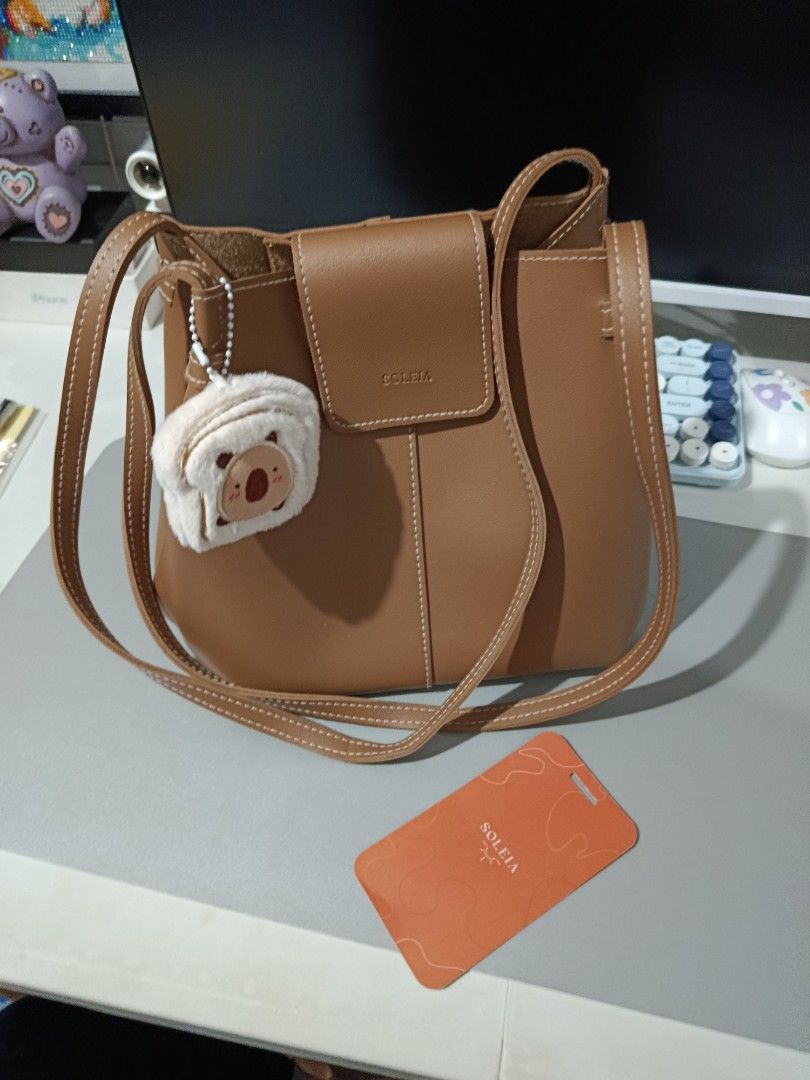 Soleia Brown Leather Shoulder Bag with Koala Charm (Category: Shoulder Bags) located in Pasay City. Submitted by pitchiiieee_ (Listing ID 1464738159, Image 3). Description: - Brand: Soleia - Material: Smooth brown leather with white contrast stitching - Closure & Straps: Front flap closure with double shoulder straps - Interior: Open pocket for small essentials - Inclusions: Fluffy koala plushie keychain and original orange brand card tag.
