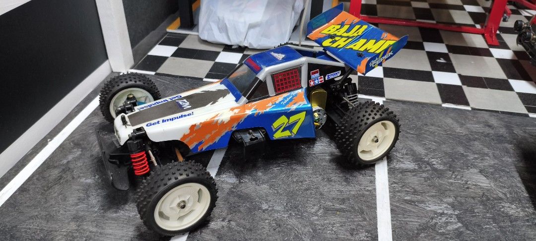 Tamiya Baja Champ RC Car (Category: Remote Control Toys) located in Caloocan City. Submitted by Outlaw (Listing ID 1464592132, Image 2). Description: - Tamiya Baja Champ remote control car - Eye-catching blue and orange livery with '27' and 'BAJA CHAMP' graphics - Shows some signs of use on body and tires, adding character - Underside view reveals chassis, motor, and servo - Ready for exciting off-road adventures - Perfect for RC car collectors a