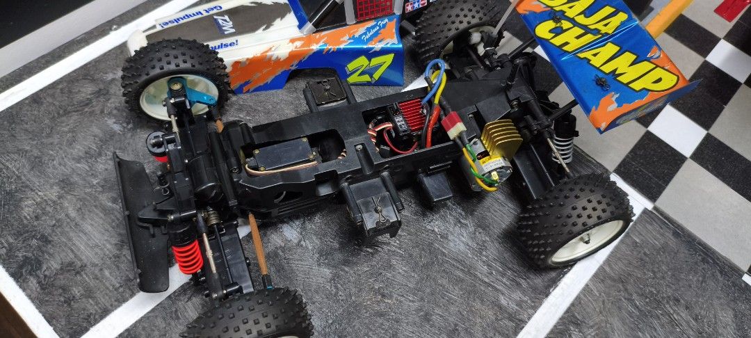 Tamiya Baja Champ RC Car (Category: Remote Control Toys) located in Caloocan City. Submitted by Outlaw (Listing ID 1464592132, Image 3). Description: - Tamiya Baja Champ remote control car - Eye-catching blue and orange livery with '27' and 'BAJA CHAMP' graphics - Shows some signs of use on body and tires, adding character - Underside view reveals chassis, motor, and servo - Ready for exciting off-road adventures - Perfect for RC car collectors a