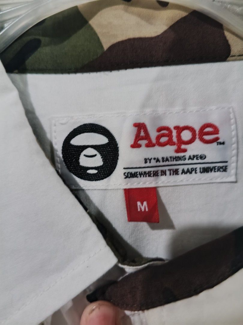 AUTHENTIC Aape By A Bathing Ape White Button-Up Shirt Medium (Category: Tshirts & Polo Shirts ) located in Cabuyao City. Submitted by full_flip0215 (Listing ID 1464901488, Image 2). Description: - Brand: Aape by A Bathing Ape - Item: White button-up shirt - Size: Medium (fits to large) Length: 28 Width : 22 - Material: 97.3% cotton, 2.7% polyurethane - Details: Embroidered "Aape Unvs" logo, bottom hem graphics, and signature camo placket tag - Co