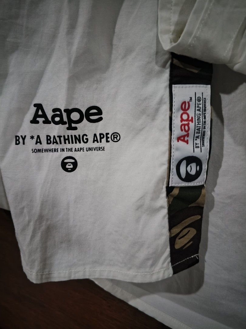 AUTHENTIC Aape By A Bathing Ape White Button-Up Shirt Medium (Category: Tshirts & Polo Shirts ) located in Cabuyao City. Submitted by full_flip0215 (Listing ID 1464901488, Image 3). Description: - Brand: Aape by A Bathing Ape - Item: White button-up shirt - Size: Medium (fits to large) Length: 28 Width : 22 - Material: 97.3% cotton, 2.7% polyurethane - Details: Embroidered "Aape Unvs" logo, bottom hem graphics, and signature camo placket tag - Co