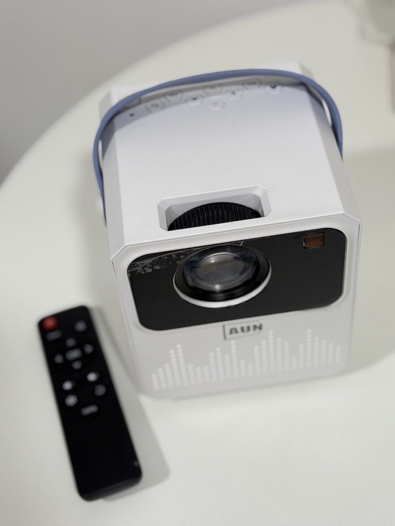 AUN Portable Mini Projector with Remote Control (Category: Projectors) located in Manila. Submitted by preshyyyyy_ (Listing ID 1464952410, Image 2). Description: - Model: Portable AUN Projector - Color: White finish with a stylish blue handle strap - Includes: Matching black remote control - Audio: Built-in speaker with a front grille pattern - Controls & Focus: Top-panel control buttons and a manual focus dial near the lens - Connectivity: Equip