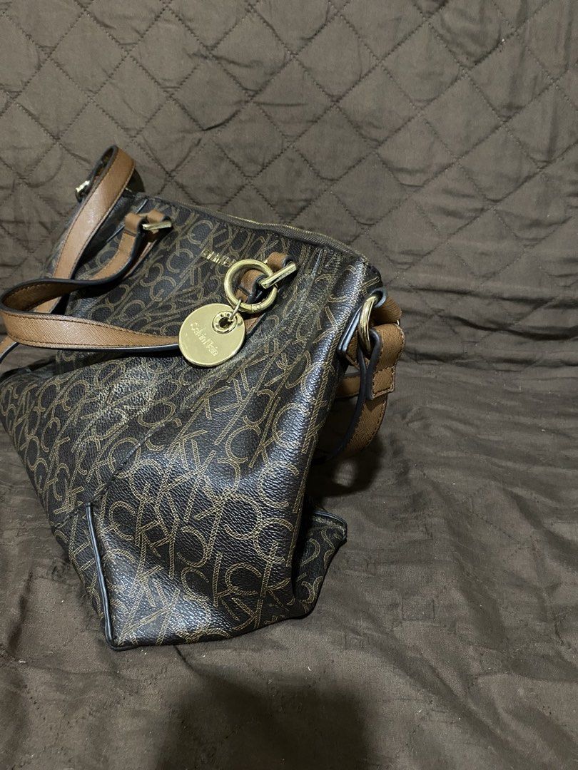 Calvin Klein monogram brown satchel shoulder bag (Category: Shoulder Bags) located in Manila. Submitted by divinemercy_29 (Listing ID 1464893087, Image 3). Description: - Brand: Calvin Klein - Design: Allover CK monogram pattern in brown with contrast tan handles - Strap: Comes with a long adjustable shoulder strap for versatile wear - Hardware: Elegant gold-tone hardware with a stylish brand charm - Closure & Interior: Secure zip-top closure wit