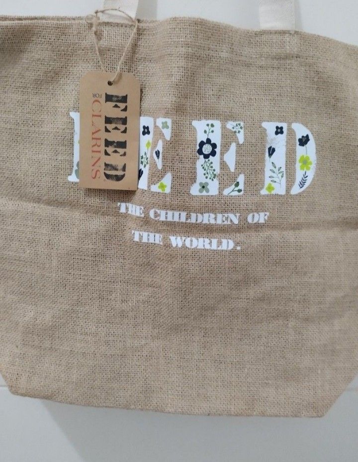 Clarins Burlap Tote Bag (Category: Tote Bags) located in Mabalacat. Submitted by uniqlover (Listing ID 1464888115, Image 3). Description: Feed for Clarins collaboration burlap tote bag. Natural jute material with sturdy cream fabric handles. Front features large white block letters filled with floral graphics and "The children of the world." text. Clarins brand tag on the side seam. Includes original cardboard hang tag attached by twine. Spacious