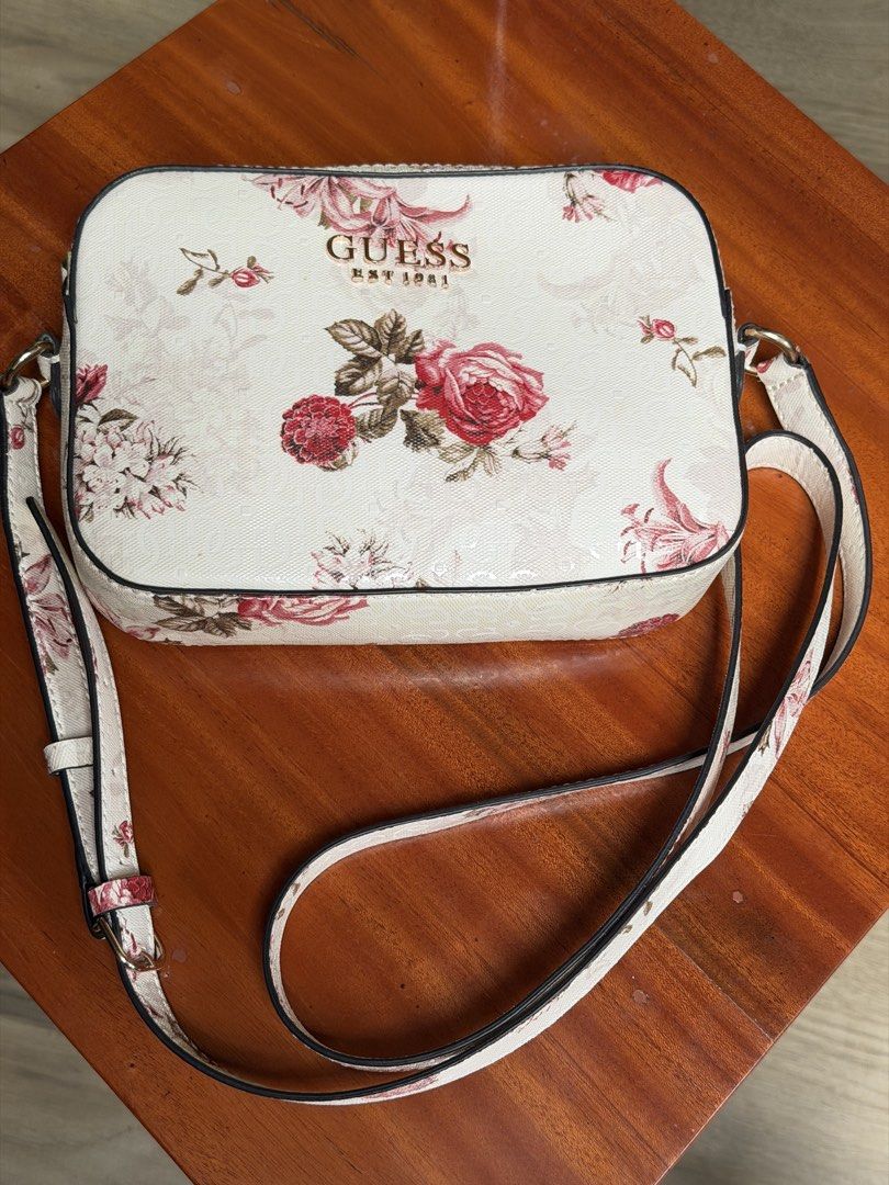 Guess Floral Crossbody Bag (Category: Cross-body Bags) located in Iloilo. Submitted by caratdeul.17 (Listing ID 1464794845, Image 1). Description: - Authentic GUESS camera bag with a white and red rose floral design - Features signature gold 'GUESS EST 1981' logo lettering on the front - Adjustable matching floral strap with clean dark piping edges - Practical rectangular shape with a secure top zip closure - Compact yet spacious enough for your 