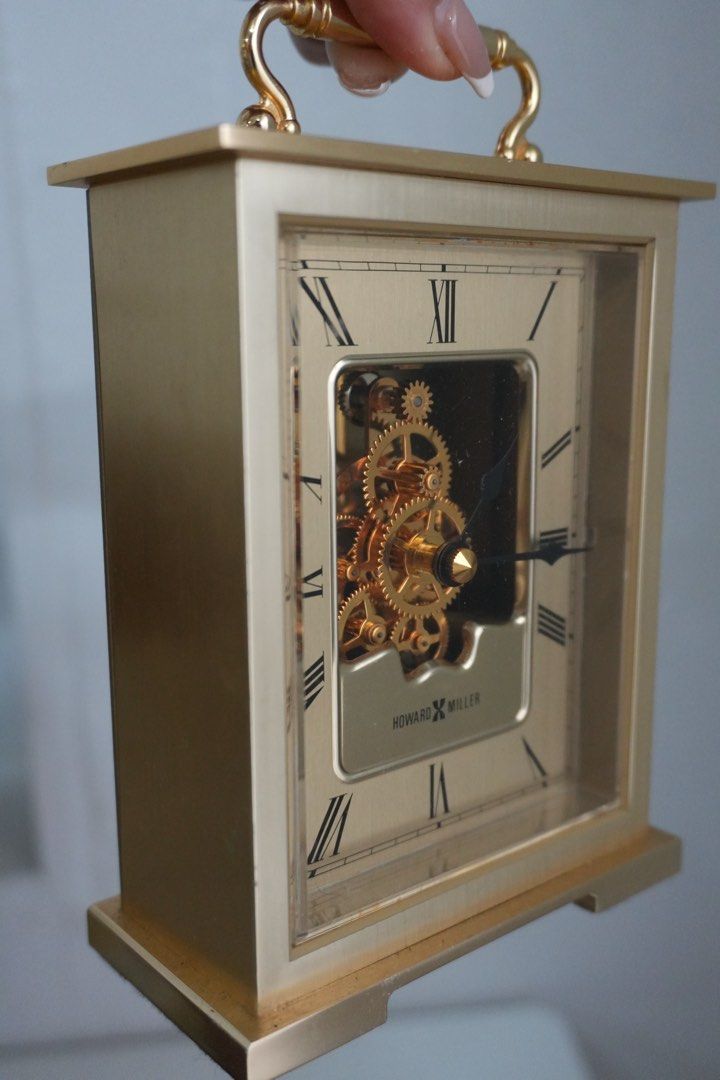 Howard Miller Skeleton Dial Carriage Tabletop Clock (Category: Clocks) located in Manila. Submitted by shopfilipina1993 (Listing ID 1464883014, Image 3). Description: - Brand: Howard Miller - Finish: gold tone with a clean polished look - Dial: Classic Roman numerals and eye-catching visible gear skeleton design - Design: Rectangular metal case, top carrying handle, flat base, and clear glass front panel - Power: Battery-operated movement - Place