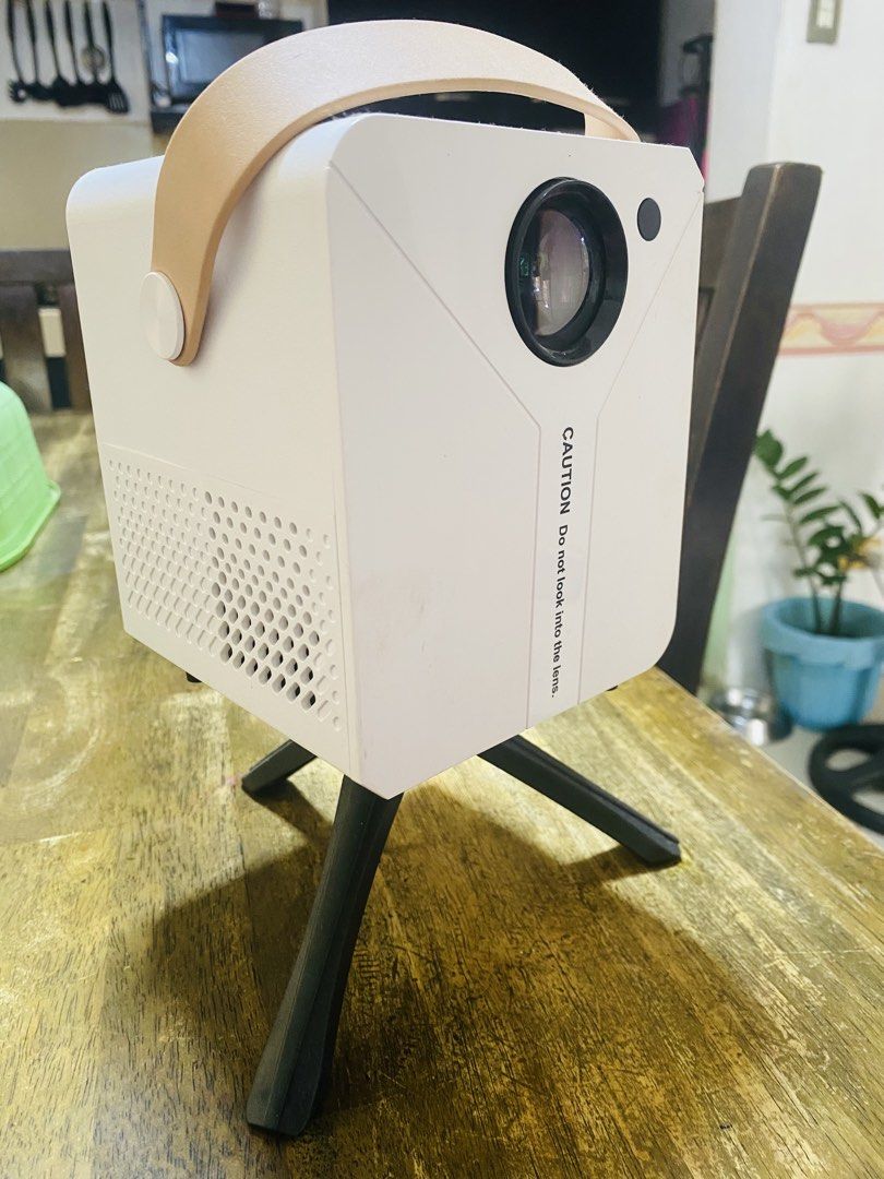 Mini Portable Projector with Tripod and Carry Strap (Category: Projectors) located in San Jose del Monte. Submitted by cityhanter007 (Listing ID 1464846083, Image 3). Description: - Compact white mini projector with a stylish tan carrying strap - Includes a sturdy black tripod stand attached to the bottom - Equipped with a power button, audio jack, USB port, and power input - Dual cooling vents on the sides and bottom to prevent overheating - Qui
