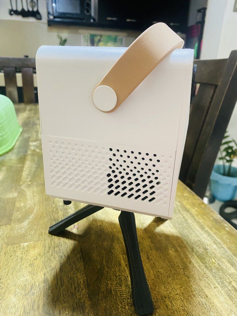 Mini Portable Projector with Tripod and Carry Strap (Category: Projectors) located in San Jose del Monte. Submitted by cityhanter007 (Listing ID 1464846083, Image 2). Description: - Compact white mini projector with a stylish tan carrying strap - Includes a sturdy black tripod stand attached to the bottom - Equipped with a power button, audio jack, USB port, and power input - Dual cooling vents on the sides and bottom to prevent overheating - Qui