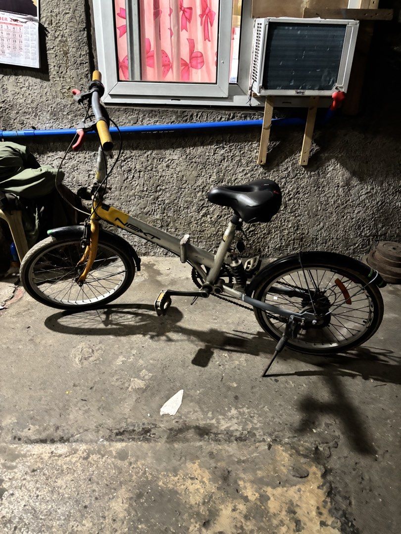 Next Folding Bike Yellow Grey (Category: Bicycles) located in Pasig City. Submitted by maanspreloved (Listing ID 1464903322, Image 1). Description: - Brand: Next Folding Bike - Color: Vibrant yellow and grey - Features: Yellow grips, red brake levers, black fenders, and rear suspension - Gears: Twist shifter on the right handlebar - Condition: Fully functional with visible rust on the chain and hardware - Extras: Includes a sturdy kickstand.