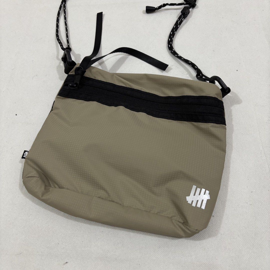 Undefeated Khaki Sling Bag with Mesh Pocket (Category: Sling Bags) located in Cavite. Submitted by itsayaaa (Listing ID 1464836894, Image 2). Description: - Undefeated crossbody bag in khaki and olive green mesh - Lightweight and durable ripstop material - Adjustable black rope shoulder strap with clips for a custom fit - Secure zippered main compartment - Front mesh pocket with snap button closure - Iconic white Undefeated logo printed near the 