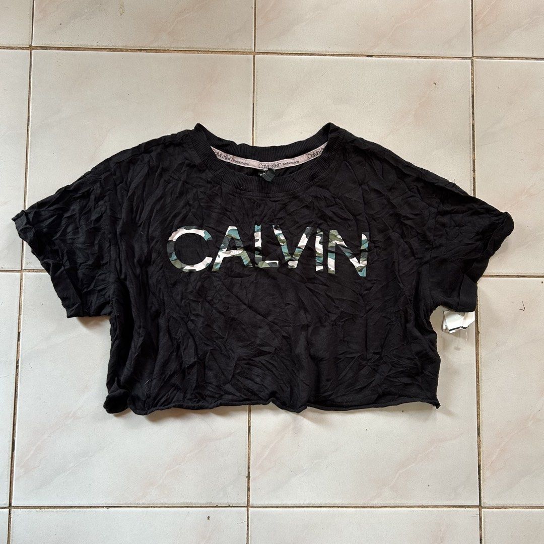 Calvin Klein Black Camo Crop Top 😍 (Category: Shirts) located in Jinoba-an. Submitted by eegiboy (Listing ID 1465001758, Image 1). Description: - Brand: Calvin Klein Performance - Size: S (Brand new with tags attached!) - Design: Bold green and white camo print spelling out 'CALVIN' across the chest - Details: Short sleeves, ribbed neckline, and small white logo near the sleeve cuff - Material: 60% cotton, 40% modal (super soft and breathable) -