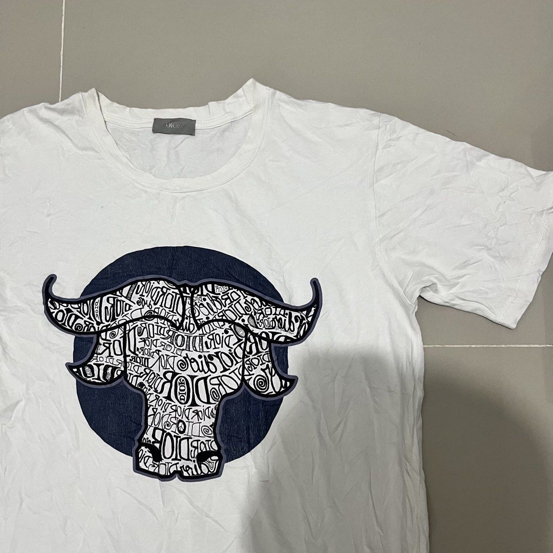 Dior Buffalo Graphic White T-Shirt (Category: Shirts) located in San Fernando. Submitted by cyrelregal27910 (Listing ID 1465124006, Image 2). Description: - Brand: Dior - Size: M (Regular Fit) - Material: 100% cotton with acrylic blend embroidery (Made in Italy) - Design: White tee featuring a blue and white buffalo graphic with intricate typographic details - Style: Crew neck, short sleeves - Condition: Clean tags and wash labels intact.
