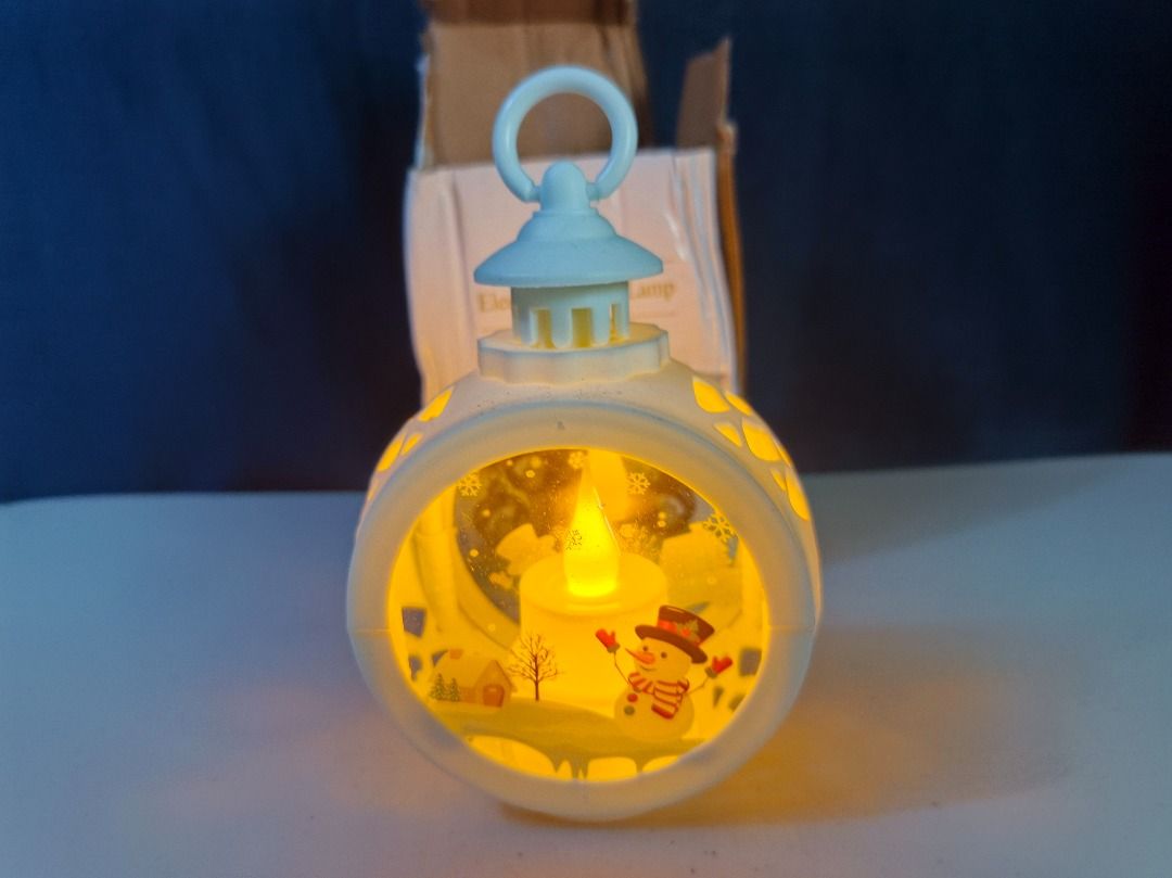 Electronic Wind Lamp Snowman Christmas LED Light (Category: Lighting) located in San Leonardo. Submitted by burgossanleonardo.12345 (Listing ID 1465051650, Image 3). Description: - White decorative electronic wind lamp with cute snowman and snowy village backdrop - Built-in LED candle provides a cozy, warm yellow light - Features a top hanging loop for versatile display options - Ideal holiday decor piece for tabletops, shelves, or hanging - Come
