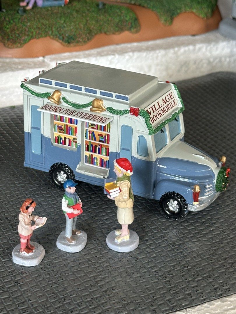 Lemax Bookmobile Christmas Village (Category: Designer & Blind Box Toys) located in Santiago. Submitted by kitalpanga (Listing ID 1465058015, Image 2). Description: - Festive blue and white bookmobile truck holiday village piece - Decorated with green garlands, bows, and bells on the roof - Side opens up to reveal display shelves filled with tiny books - Includes 3 adorable miniature figures: a woman holding books, a boy, and a small child readin