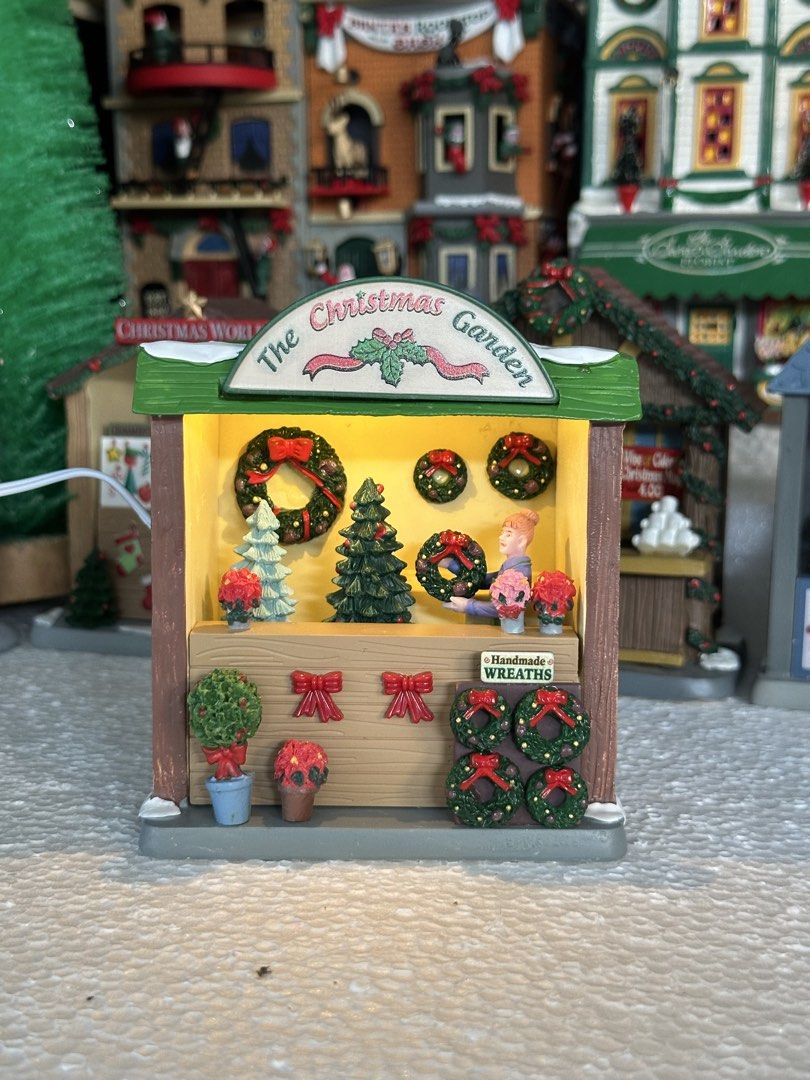 Lemax The Christmas Garden (Category: Figurines & Statues) located in Santiago. Submitted by kitalpanga (Listing ID 1465062838, Image 3). Description: Lighted. Adaptor not included - Miniature holiday market stall named 'The Christmas Garden' - Includes a shopkeeper figure holding a green wreath behind the counter - Features detailed handmade wreaths, small potted poinsettias, and decorated evergreen trees - Designed with a festive green roof and