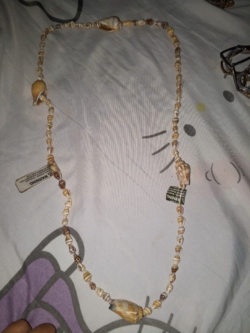 Natural seashell beaded necklace with warning tags (Category: Necklaces) located in Magalang. Submitted by zeiansoria64090 (Listing ID 1464980387, Image 2). Description: - Long seashell necklace that hangs down the chest - Features a mix of various brown and white shells - Accented with multiple larger shell accents throughout the strand - Perfect earthy beach-style accessory for summer outfits - Warning: Contains small parts, choking hazard labe