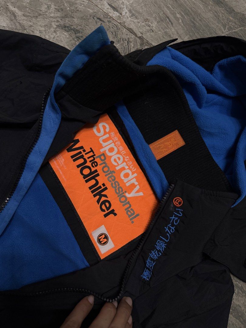 Superdry The Windhiker Jacket Black Blue Size M (Category: Coats, Jackets and Outerwear) located in Alaminos. Submitted by outfitcheck.ph (Listing ID 1465109768, Image 2). Description: - Brand: Superdry The Windhiker - Color: Black with bright blue inner fleece lining - Size: Medium (M) - Features: Full zip front, zippered chest pocket, and adjustable hood toggles - Details: Japanese character embroidery on storm flap and back right shoulder, plu
